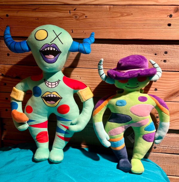 wo colorful plush toys with unique, playful designs sit against a wooden background. One toy features a green body, blue horns, and expressive multi-mouth details, while the other has a green body, white horns, a purple hat, and bold red eyes. Their vibrant patches and stripes add a striking visual contrast, perfect for collectors or fans of artistic plush designs. These plush companions come with an interactive activity book and a mysterious alien alphabet, inviting imaginative play and decoding adventures. creature speak alien plushie